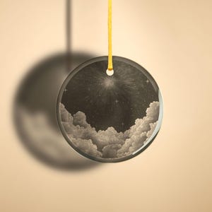 Op de afbeelding: Een ronde glazen ornament met een zwart-wit afbeelding van een sterrenhemel met wolken. De ornament hangt aan een geel lint.