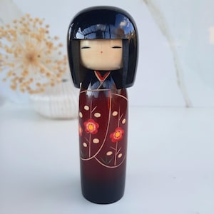 Puede incluir: Una muñeca kokeshi japonesa tradicional con cabello negro, un kimono estampado en rojo y blanco y una cara pintada. La muñeca está hecha de madera y tiene una forma redondeada.
