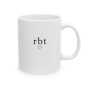 May include: A white ceramic mug featuring the lowercase black text rbt centered on the front, positioned directly above a small, simple black outline of a heart. The mug has a smooth, cylindrical body and a curved handle on the right side.