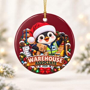 May include: A round maroon Christmas ornament featuring a cartoon penguin wearing a Santa hat and a high-visibility vest. The penguin holds cardboard boxes near a pallet jack, a clipboard, and shelves. The text reads WAREHOUSE ASSOCIATE 2026.