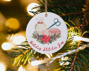 May include: White ceramic Christmas ornament with a watercolor illustration of a sewing kit, a spool of thread, and a poinsettia. The ornament has the text "Holiday Stitches Joy" written around the image.