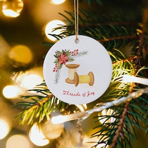 May include: A white ceramic Christmas ornament with a red poinsettia and green holly leaves. Two wooden spools of thread are shown with the text "Threads of Joy".
