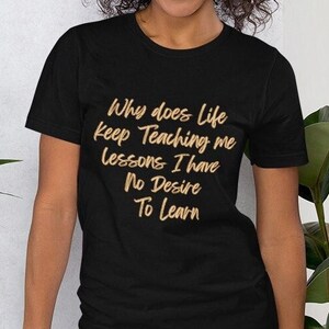 May include: Black t-shirt with the text "Why does life keep teaching me lessons I have no desire to learn" in a gold script. The shirt is paired with blue jeans.