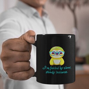 Puede incluir: Taza de café negra con un perezoso de dibujos animados con gafas y sombrero. La taza tiene el texto "I'm fueled by slow, steady laziness" en escritura turquesa. El perezoso es amarillo con gafas azules.