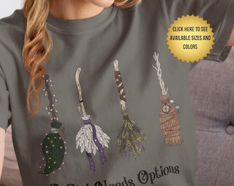 Könnte beinhalten: Graues T-Shirt mit vier illustrierten Hexenbesen in verschiedenen Designs. Der Text "A Girl Needs Options" ist unter den Besen aufgedruckt. Eine goldene Sternform in der oberen rechten Ecke zeigt "Hier klicken, um verfügbare Größen und Farben zu sehen."