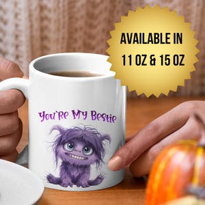 Puede incluir: Taza de cerámica blanca con el texto "You're My Bestie" y una ilustración de un monstruo morado. La taza está sostenida por una persona, con un plato de galletas y una calabaza en el fondo.