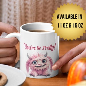 Puede incluir: Taza de cerámica blanca con un monstruo rosa y peludo y el texto "You're So Pretty!". La taza contiene una bebida oscura y está disponible en tamaños de 312 ml y 426 ml. Un plato con galletas y calabazas está en el fondo.