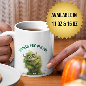 Puede incluir: Taza de cerámica blanca con el texto "I'm your HUG in a MUG" y una ilustración de un monstruo verde. La taza está sostenida por una persona. Un plato con galletas y una calabaza están en el fondo.