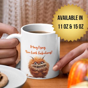 Puede incluir: Taza de cerámica blanca con una ilustración de monstruo de dibujos animados y el texto "May I say, You Look Fabulous!". La taza está disponible en tamaños de 312 ml y 425 ml.