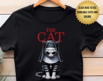 May include: A black short-sleeve t-shirt featuring a graphic of a cat dressed as a nun with a cross necklace. Above the cat, the text reads THE CAT in red capital letters. A gold starburst badge in the top right corner says CLICK HERE TO SEE AVAILABLE SIZES AND COLORS.