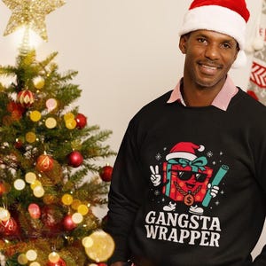 Puede incluir: Una sudadera negra con un gráfico navideño que dice "Gangsta Wrapper". El gráfico presenta una caja de regalo con gafas de sol y un gorro de Papá Noel. Un árbol de Navidad y medias están en el fondo.