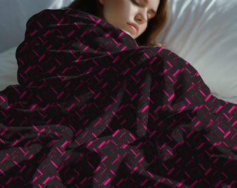 Può includere: Una coperta nera con un motivo ripetuto di forme rettangolari rosa acceso. La coperta è drappeggiata su qualcuno a letto, con il cuscino bianco e la testiera visibili sullo sfondo. La coperta sembra morbida e accogliente.