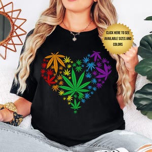 May include: Black t-shirt with a heart-shaped design composed of cannabis leaves in rainbow colours. The text "CLICK HERE TO SEE AVAILABLE SIZES AND COLORS" is also visible on the shirt.