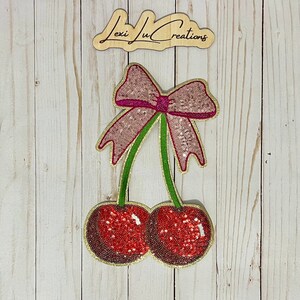 May include: A pink and red sequined bow with two red cherries with gold outlines. The cherries have green stems.