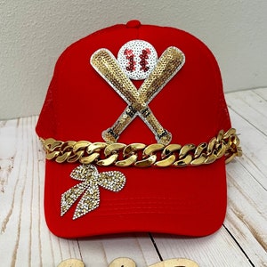 May include: Red mesh baseball cap with a gold chain and a gold sequined baseball and bats design. The cap also has a gold sequined bow on the side.