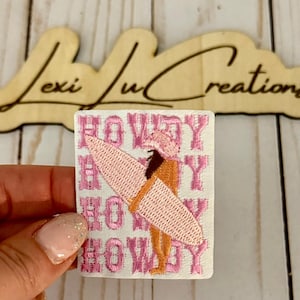 Puede incluir: Parche bordado con una tabla de surf rosa y la palabra "HOWDY" repetida en rosa sobre un fondo blanco. El parche también representa a una persona con un atuendo naranja. El parche está sostenido por una mano con esmalte de uñas rosa brillante.
