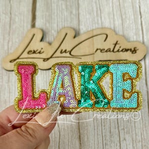 May include: A decorative patch featuring the word LAKE in block letters. Each letter is covered in sequins with a gold border. The L is pink, the A is light purple, the K is green, and the E is light blue. A wooden sign with the text Lexi Lu Creations is behind it.