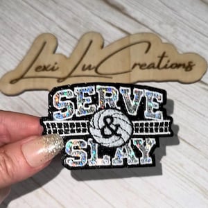 May include: A black patch with silver holographic lettering that reads SERVE & SLAY, featuring a central white volleyball graphic and a horizontal net pattern. The text and volleyball are outlined in white, creating a high-contrast design.