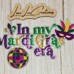 May include: Wooden sign with the text "Lexi Lu Creations" above a collection of sequined Mardi Gras-themed decorations. The decorations include a mask, a disco ball, and the phrase "In my Mardi Gras era."