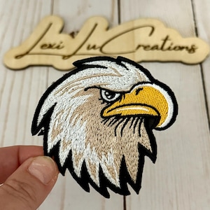 May include: Embroidered patch of an eagle's head, showcasing a detailed design with a black border, tan and white feathers, and a yellow beak. The patch is set against a light wooden surface, with the text "Lexi Ju Creations" in a wooden cutout.