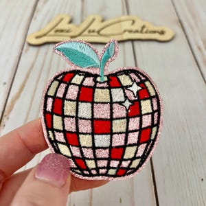 May include: A pink and red glitter iron-on patch shaped like an apple with a disco ball design. The apple has a green leaf and white stars on the side.
