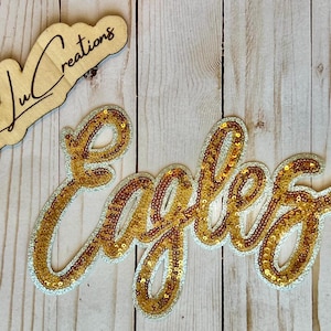 May include: A wooden sign with the text "Lexi Ju Creations" and a gold sequined "Eagles" patch. The patch has a silver border and is on a light wood background.