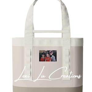 May include: A beige tote bag with a cream-colored top and handles. The bottom of the bag is a darker beige. A small photo is centered on the front. The words "Lexi Lu Creations" are written in a cursive font across the front of the bag.