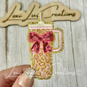 May include: An embroidered patch shaped like a tumbler with a straw, featuring a pink and gold leopard print pattern, a gold glitter border, a light pink handle, and a dark pink sequined bow centered on the front of the cup design.