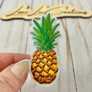 May include: Embroidered pineapple patch with a detailed design. The patch features a yellow and brown pineapple body with green leaves. The patch is held by a hand against a light wood background.