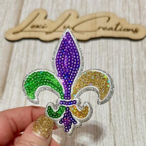 May include: A sequined fleur-de-lis in green, purple, and gold with a silver glitter outline. The design is held by a hand with a gold glitter nail. A wooden sign with the text "Lexi Lux Creations" is in the background.