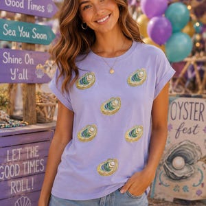 May include: A light purple short-sleeve t-shirt featuring five embroidered oyster shell motifs with pearl accents, paired with light blue denim shorts with a frayed hem. The shirt has a crew neckline and rolled sleeves, showcasing a casual design.