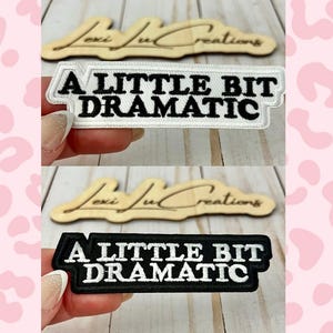 Op de afbeelding: Twee geborduurde patches met de tekst "A LITTLE BIT DRAMATIC" in witte blokletters. Eén patch heeft een zwarte rand en achtergrond, de andere een witte rand en achtergrond. De patches worden getoond op een houten oppervlak.