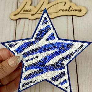 May include: A blue star-shaped sequin patch with white and blue stripes. The patch has a dark blue border and is on a light-colored wooden surface. The text "Lexi Lu Creations" is visible in the background.