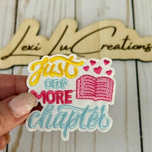 May include: Embroidered patch with the text "Just One More Chapter" in yellow, pink, and blue. The patch features a pink book with hearts above it. The patch is held in a hand, with a wooden sign in the background.