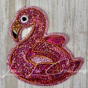 May include: A pink glitter flamingo patch with gold trim. The flamingo has a black eye, a gold beak, and a white area around the beak. The patch is in a relaxed pose, with its wings slightly open.