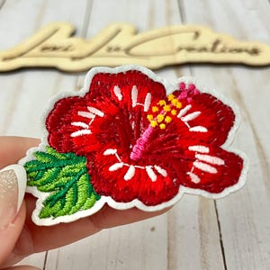 May include: Embroidered red hibiscus flower patch with white accents, green leaves, and yellow stamens. The patch is on a white background. The patch is held by a person. The wooden sign in the background says "Lexi Lu Creations".