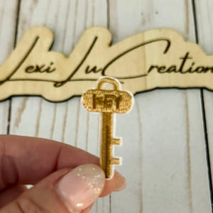Puede incluir: Un parche dorado en forma de llave con la palabra "KEY" bordada en la parte superior. El parche se sostiene frente a un letrero de madera que dice "Lexi Lu Creations". El fondo es una superficie de madera de color claro.