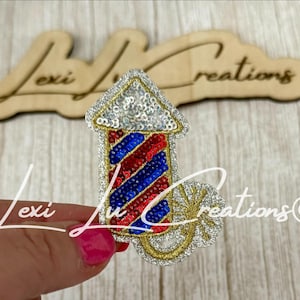 May include: A sequined firework pin with red, white, and blue stripes. The pin has a gold-colored outline and a silver sequined top. The pin is held in a hand with pink nail polish. The background includes wooden cutouts with the text "Lexi Lu Creations".