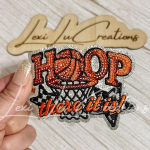 May include: A sequined patch featuring a basketball graphic and the text "HOOP there it is!" in orange and silver. The design includes a basketball hoop net pattern, with orange sequins forming the letters and silver sequins as the background.