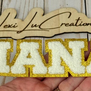 May include: A gold glitter and white felt "NANA" patch. The letters are white with a gold glitter border. A wooden sign with the text "Lexi Ju Creations" is above the patch.