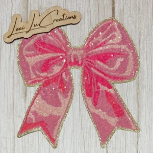 May include: A large, pink sequined bow with gold trim. The bow has a layered design with lighter pink accents. A wooden sign with the text "Lexi Lu Creations" is above the bow.