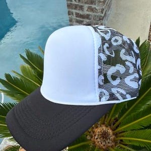 May include: A trucker hat featuring a solid white foam front panel, a black curved brim with stitched detailing, and a black and white mesh back panel decorated with a repeating leopard print pattern across the side and rear sections.