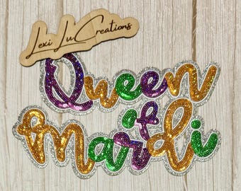May include: A wooden sign with the text "Lexi Lu Creations" above a sequined "Queen of Mardi" design. The design features gold, green, purple, and silver glitter accents, perfect for Mardi Gras celebrations.