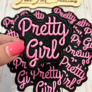 May include: Black embroidered patch with the words "Pretty Girl" in pink script. The patch has a black border and is surrounded by other similar patches. A wooden sign with the text "Lexi Ju Creations" is in the background.