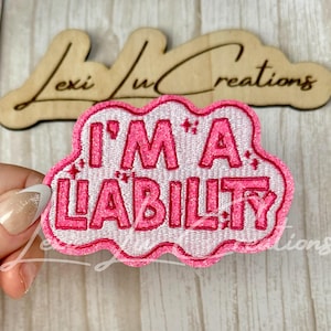 May include: A cloud-shaped embroidered patch with a white background and a bright pink border. The text "I'M A LIABILITY" is stitched in bold, glittery pink letters across the center, accented by small embroidered star shapes.