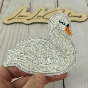 May include: A white sequined swan patch with a silver glitter outline. The swan has an orange beak, black eyes, and a curved neck. The patch is held in a hand, with a wooden sign in the background that reads "Lexi Jul Creations".