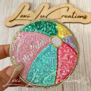 May include: A circular embroidered patch featuring a beach ball design covered in multi-colored sequins. The segments are colored in shades of green, pink, yellow, and purple, with a silver center circle and a silver glitter border trim.
