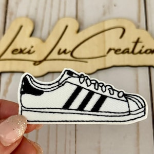 Pode incluir: Um patch de tênis branco e preto com três listras pretas na lateral. O patch está na frente de uma placa de madeira que diz "Lexi Lu Creations".