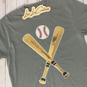 May include: A gray t-shirt with a gold sequined baseball bat and ball design. The design features two bats crossed over each other with a baseball in the center.