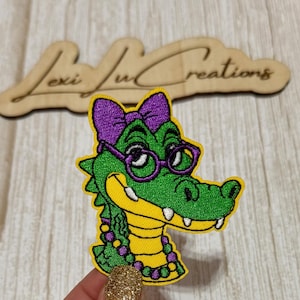 May include: Embroidered patch featuring a green alligator wearing purple glasses and a bow. The alligator has a yellow and purple necklace and is outlined in yellow. The patch has the text "Lexi Ju Creations" in a wooden cutout.